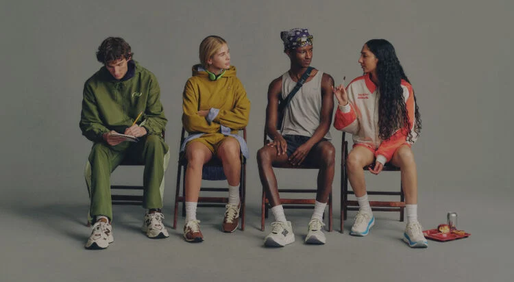Four people seated side-by-side on simple wooden chairs against a plain, neutral background, arranged in a horizontal line.